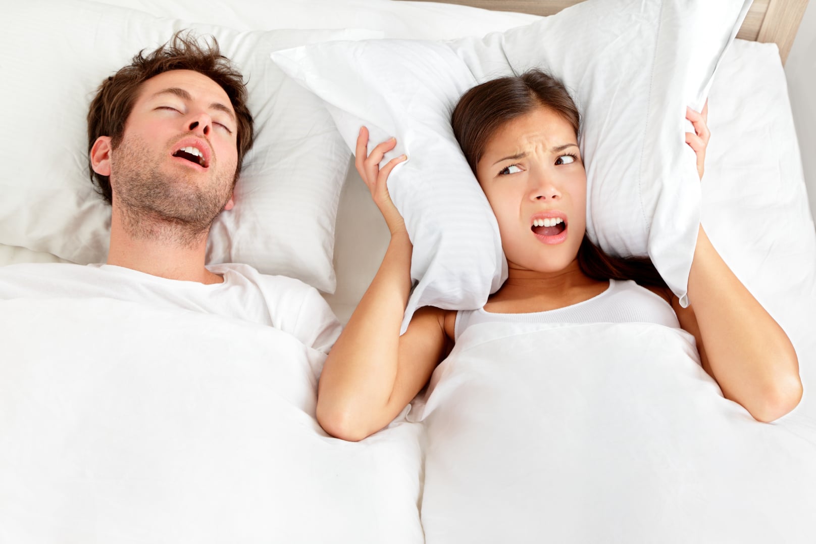 A woman and man in a bed. The woman is covering her ears with a pillow while the man snores